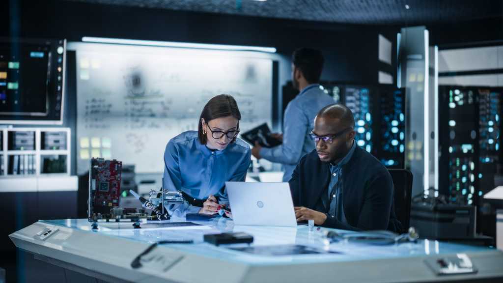 Why standardizing workplace technology is the next competitive advantage for CIOs Diverse Group of Industrial Robotics Specialists Gathered Around a Table, Using Laptop