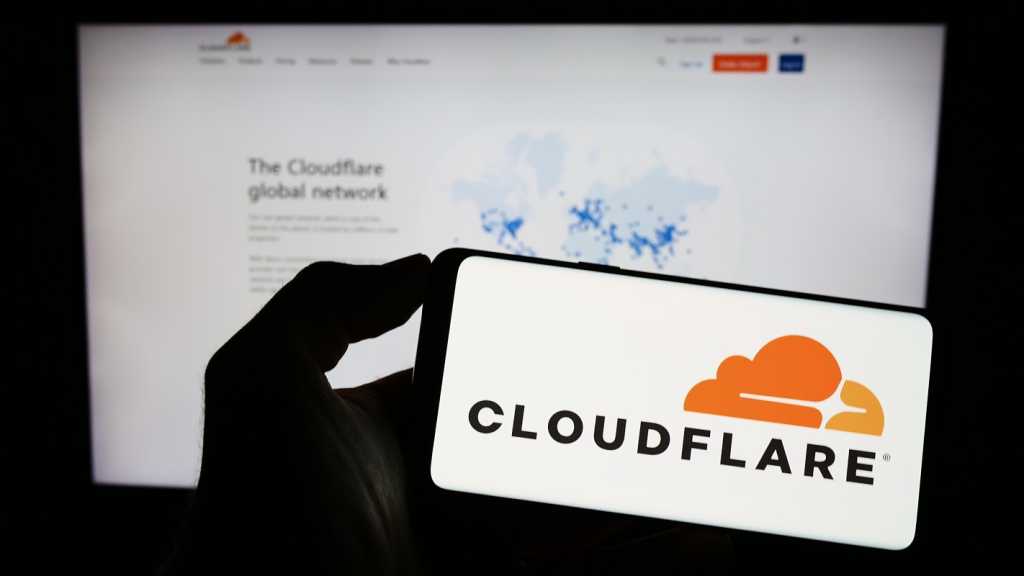 Cloudflare launches Dynamic Workers for AI agent execution A photograph of a person holding a smartphone with the Cloudflare logo displayed. Behind the phone is a blurred monitor displaying the Cloudflare website.,