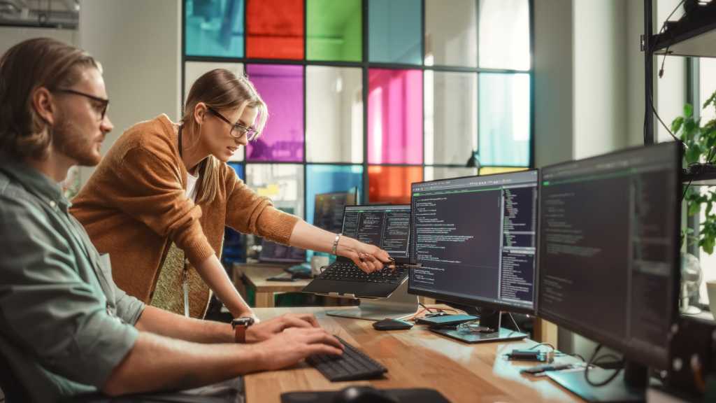 AWS targets software release bottlenecks with DevOps Agent update Female Senior Software Engineer Gives Advice to Male Junior Developer, Pointing at Desktop Computer Display With Code. Caucasian Woman Offers New Features, Helps Fixing Bugs in Modern SaaS Platform.